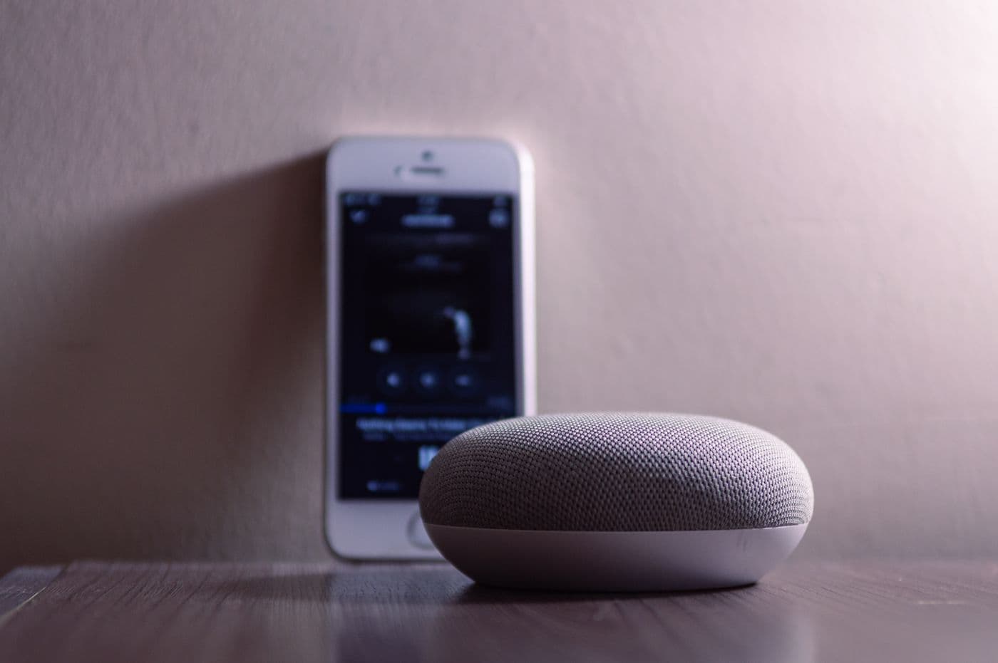 Google Home smart speaker beside a smartphone on a wooden table — voice and app control for connected home devices