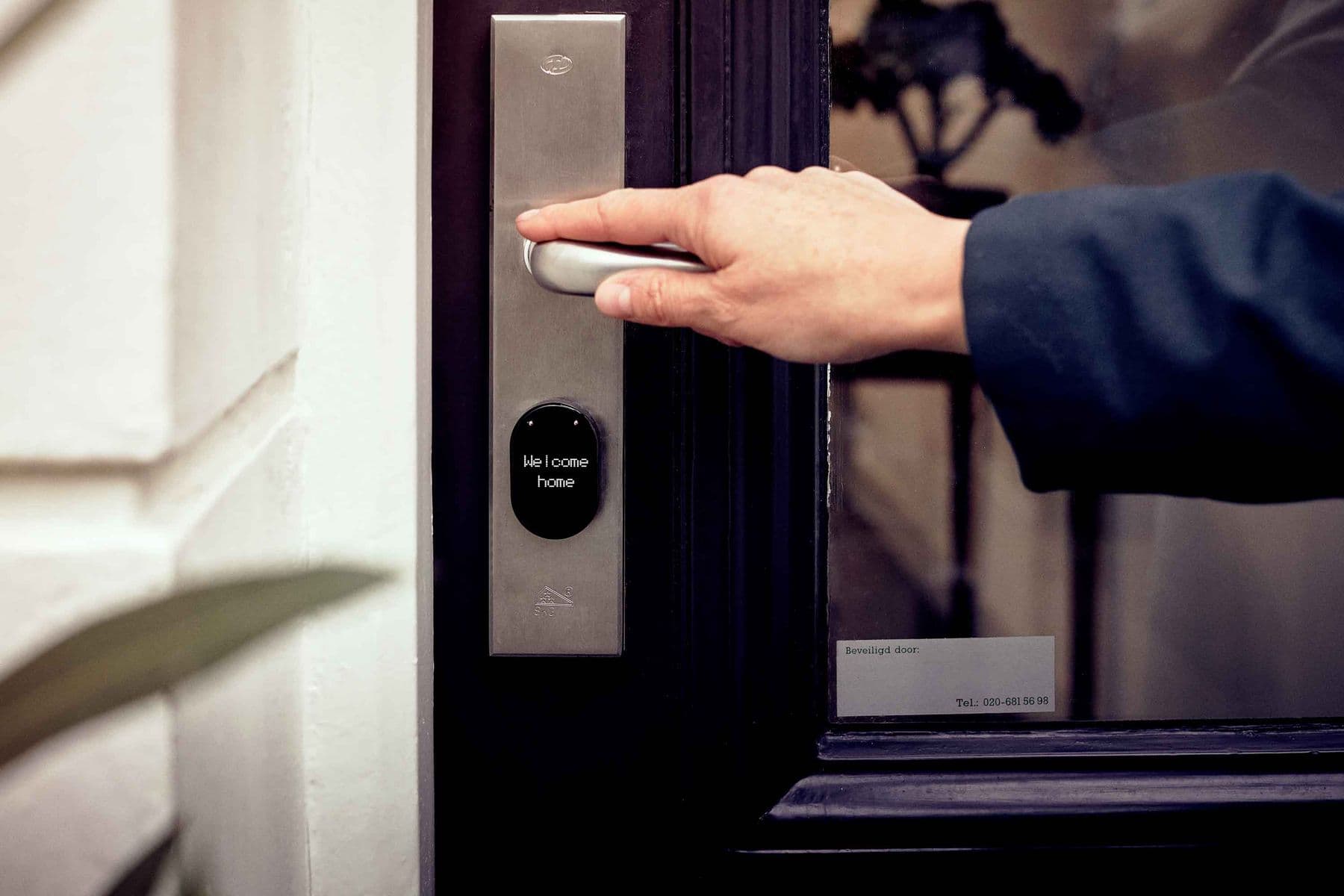 A hand opening a front door fitted with a smart lock that displays a welcome message